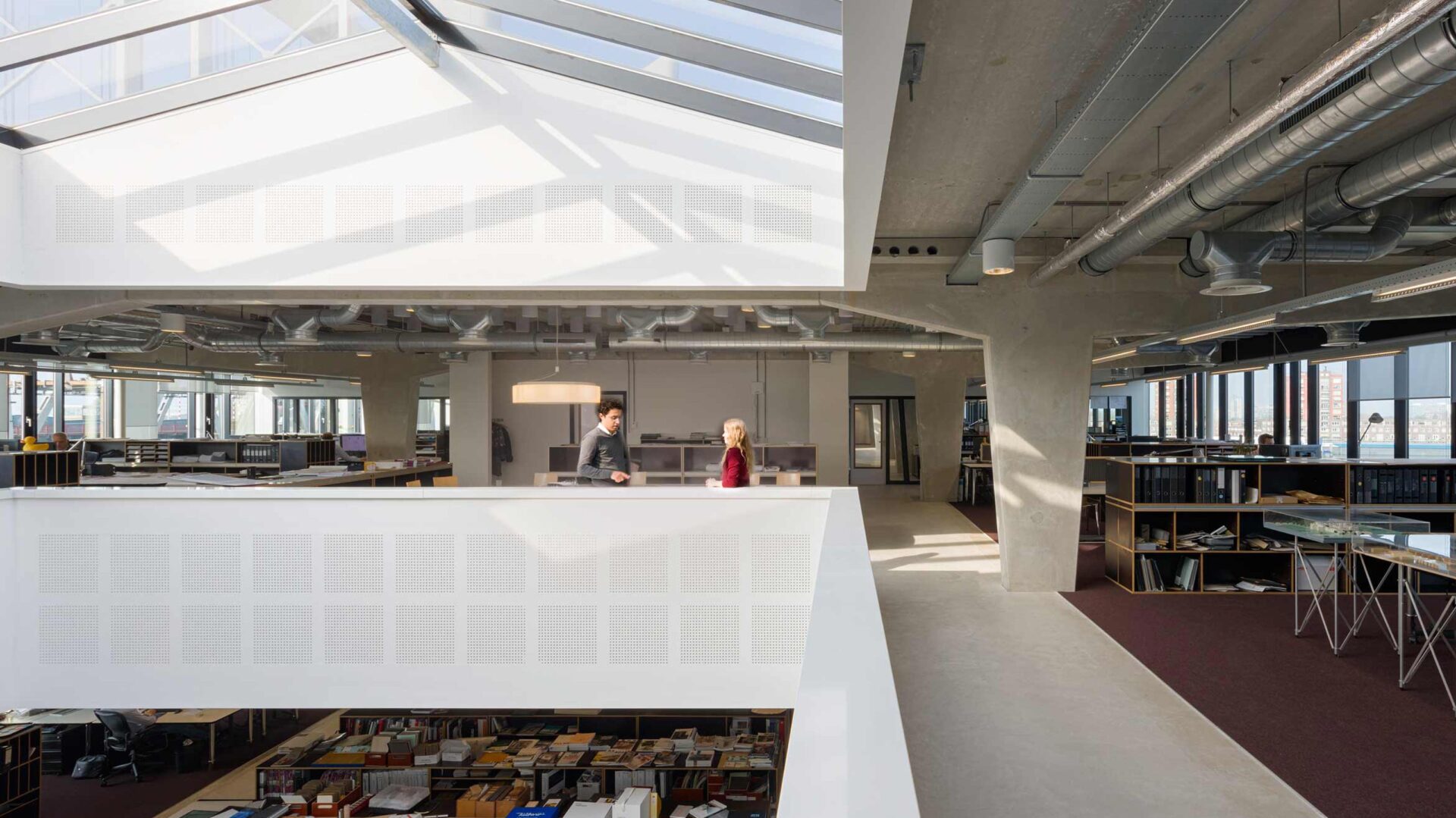 Open kantoorruimte met twee mensen bij een vide, veel daglicht en zichtbare ventilatiebuizen aan het plafond